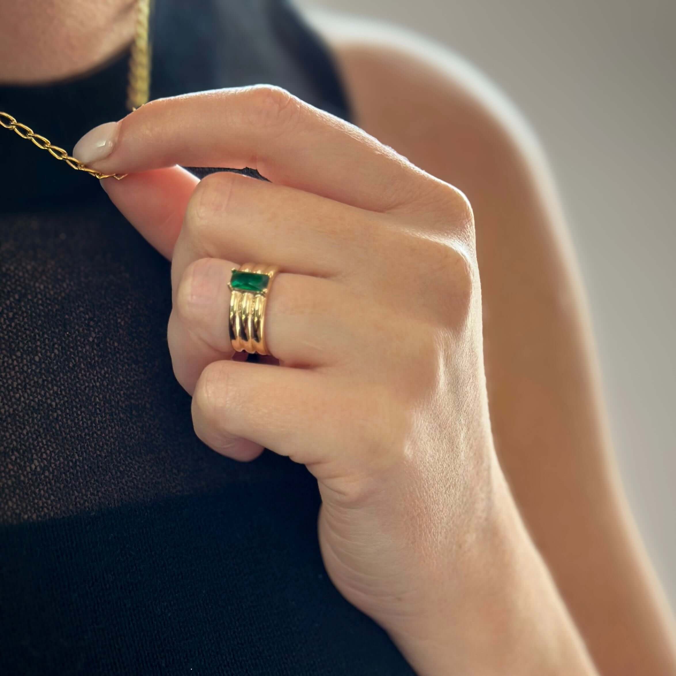 Close-up of a person's hand holding a gold necklace, wearing an Envy Emerald Ring with a green gemstone, and a sleeveless black top.