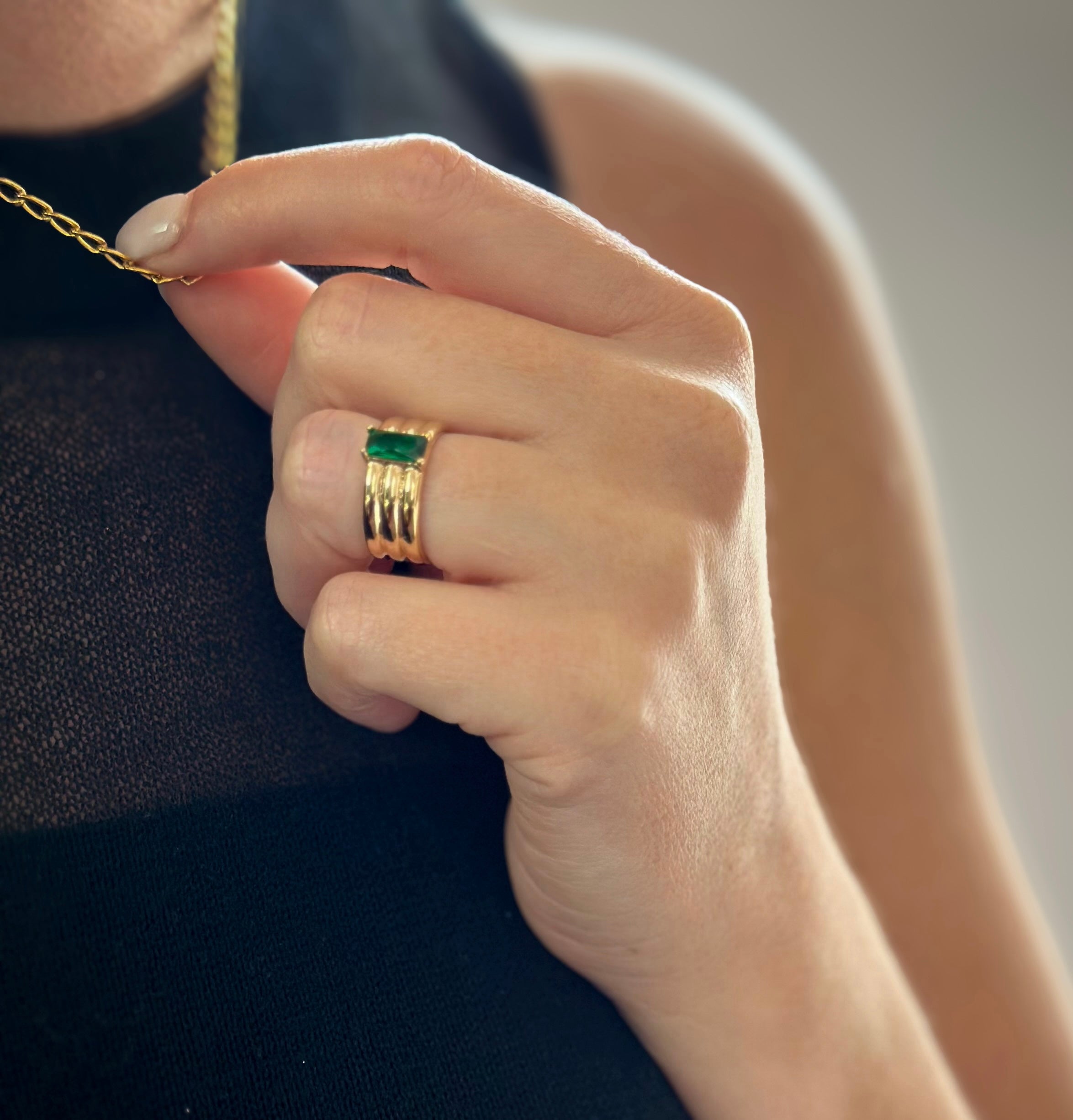 Close-up of a hand with an emerald ring holding a gold necklace against a sheer black top.