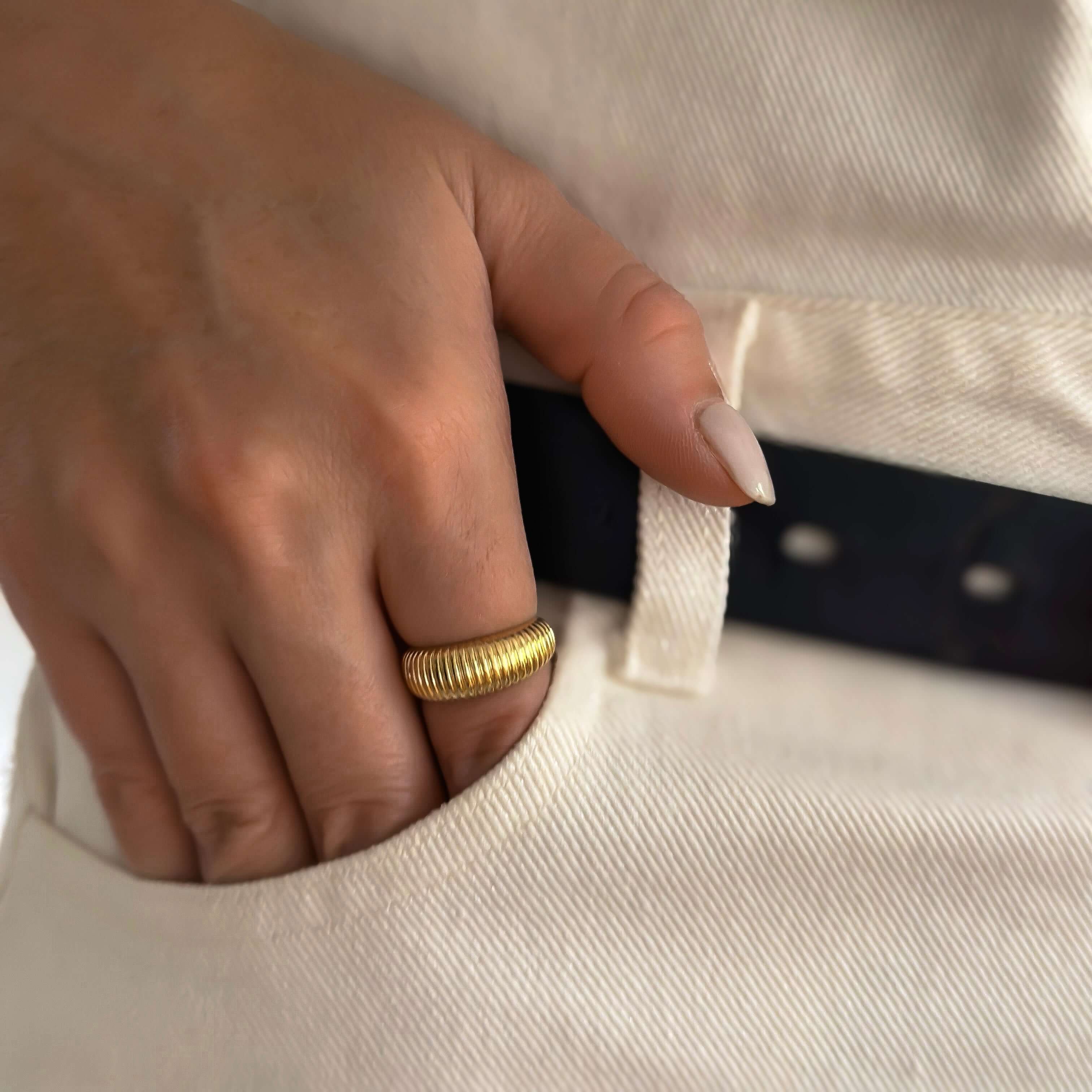 Close-up of a hand adorned with the Venice Ring, showcasing its distinctive texture and 18k gold plating. The thumb is tucked into the pocket of white pants, with a black belt also visible.