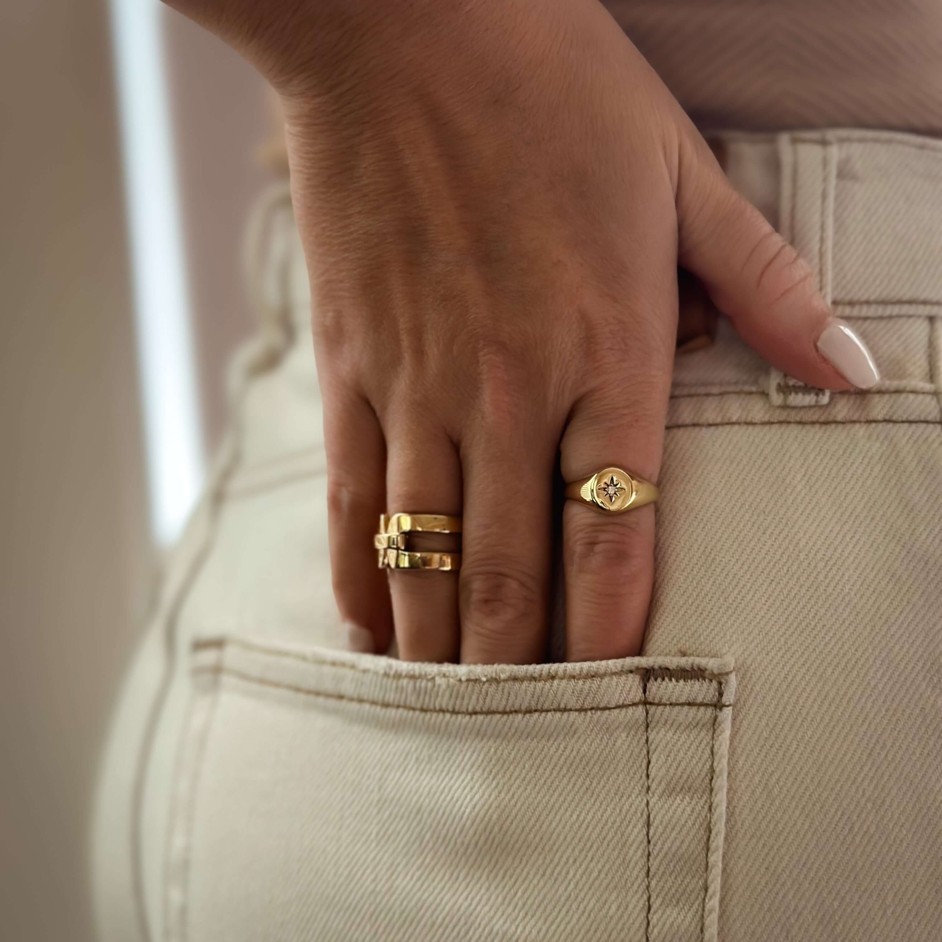 Close-up of a hand showcasing the Star Gazer Signet Ring and other gold accessories, tucked elegantly into the back pocket of light-colored pants—an epitome of celestial chic and quality craftsmanship.