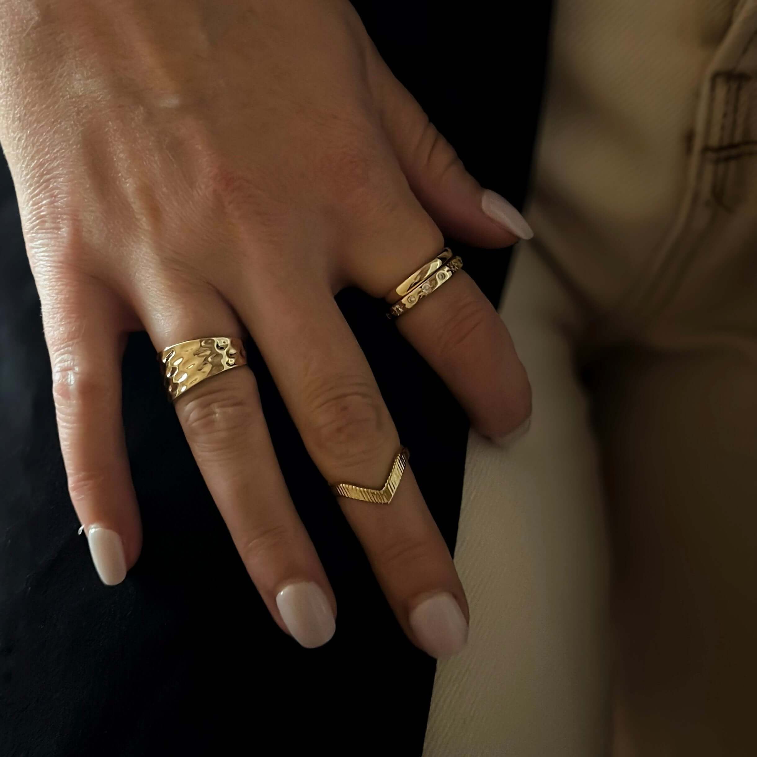 A hand with manicured nails flaunts three stylish gold rings, including an 18k gold-plated brass band and the elegant Josie Hammered Ring, adorning the middle and ring fingers.