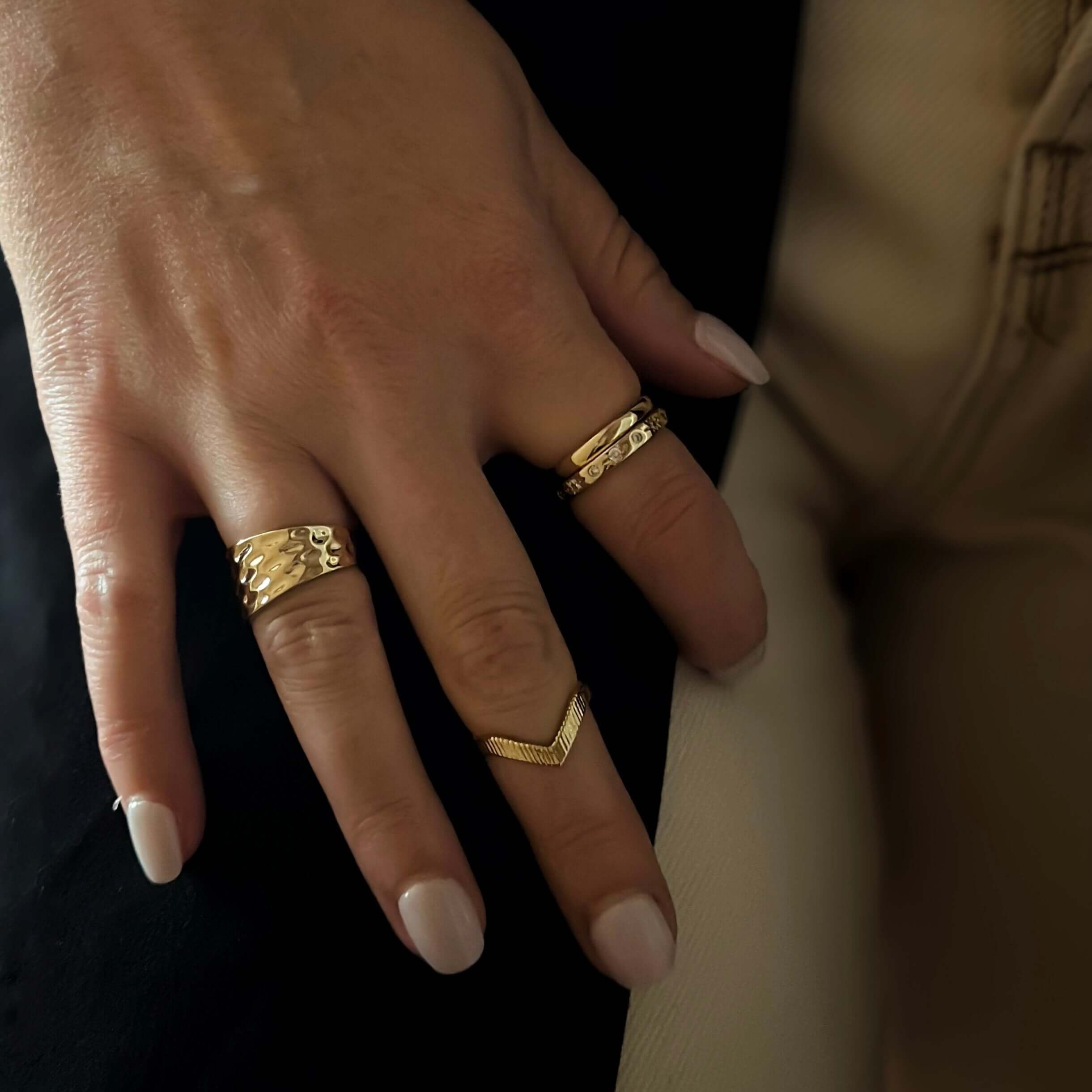 A hand with manicured nails adorned with three Essential Stacking Rings, showcasing minimalist jewelry against a dark background. The stacking rings glisten with 18k gold plating.