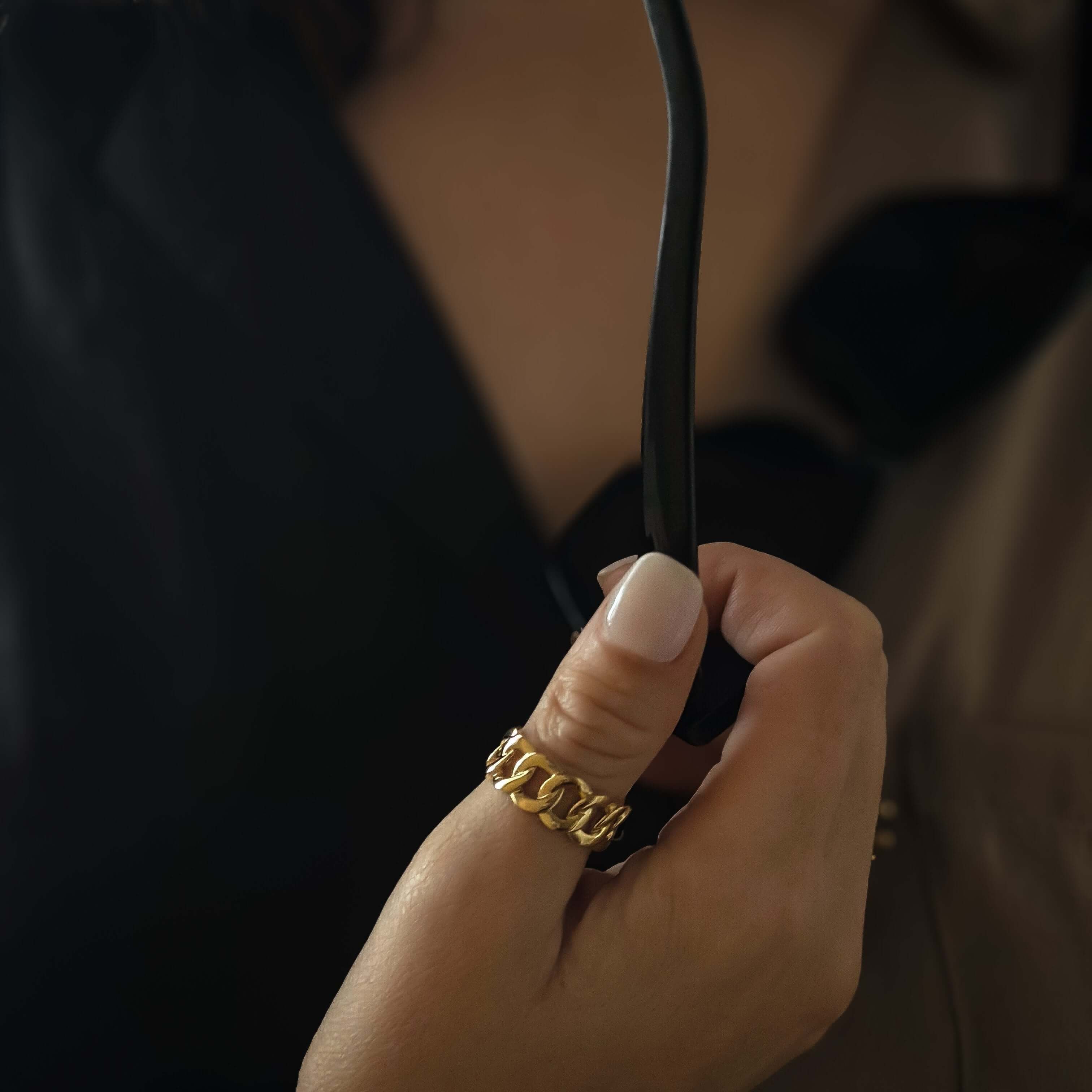 A close-up shot showcases a hand holding black sunglasses, adorned with the fashion-forward Bold Chain Ring featuring 18k gold plating, set against a dark, blurred background.