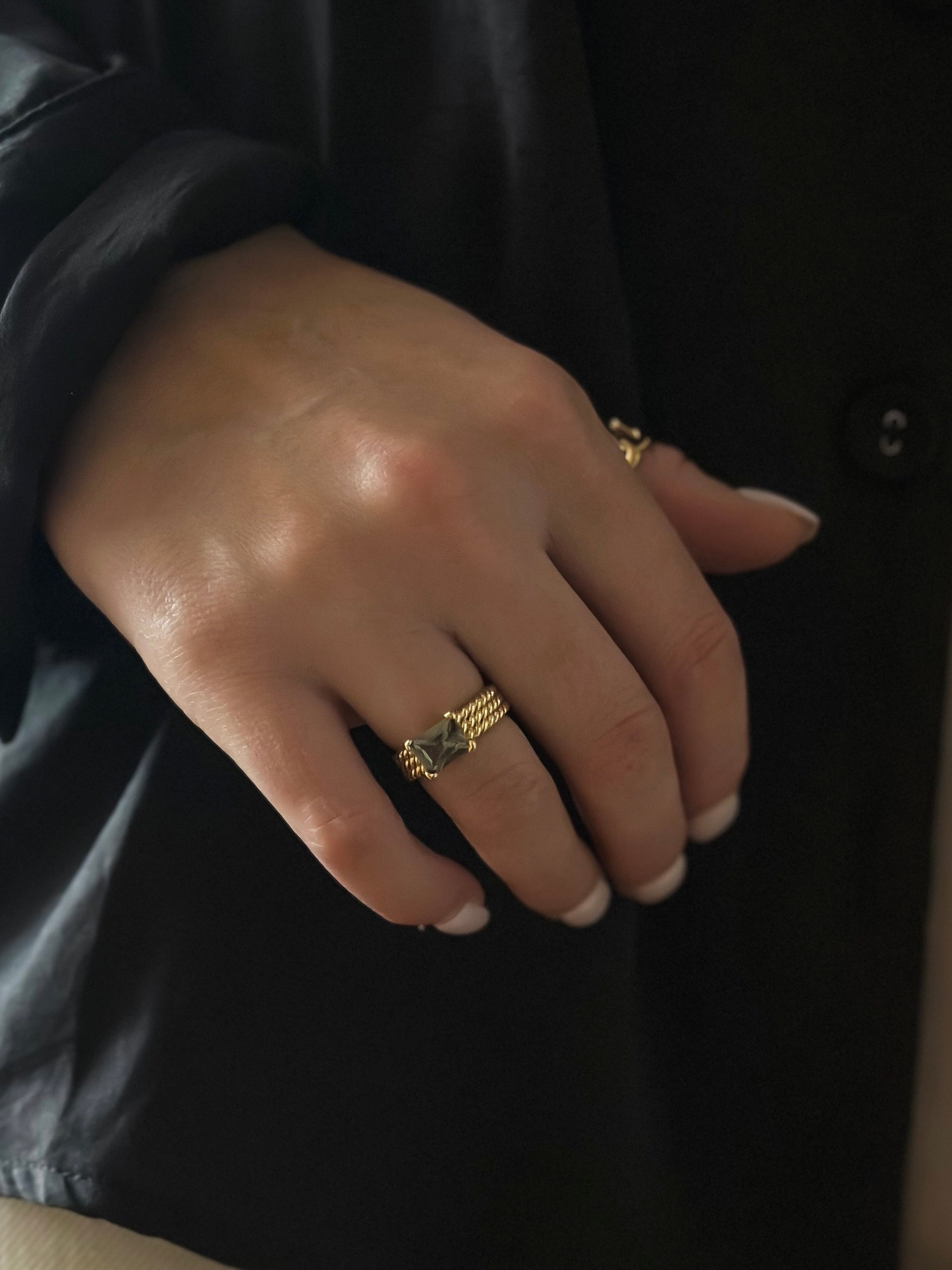 Close-up of a hand wearing gold rings, including one with a green gemstone, against a dark fabric background.