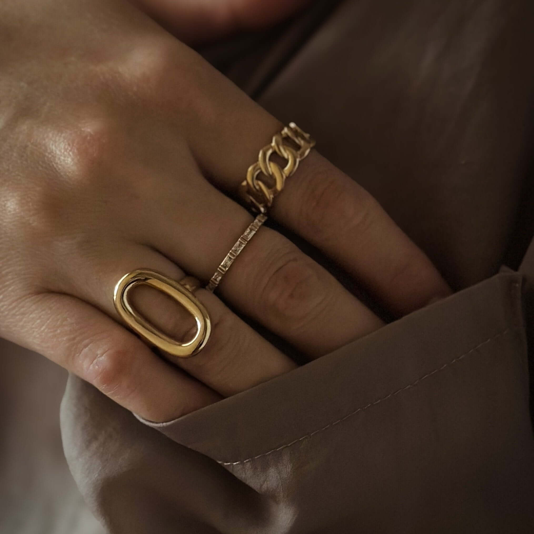 A hand adorned with luxury jewelry, including the Oval Statement Ring and two other uniquely designed gold rings, rests against a dark fabric background.