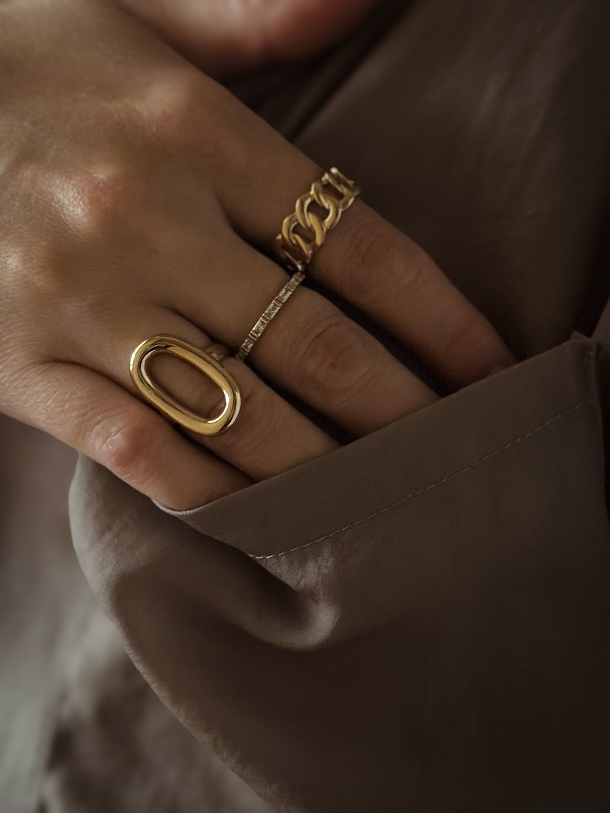 A hand adorned with three different gold rings rests on a brown fabric.