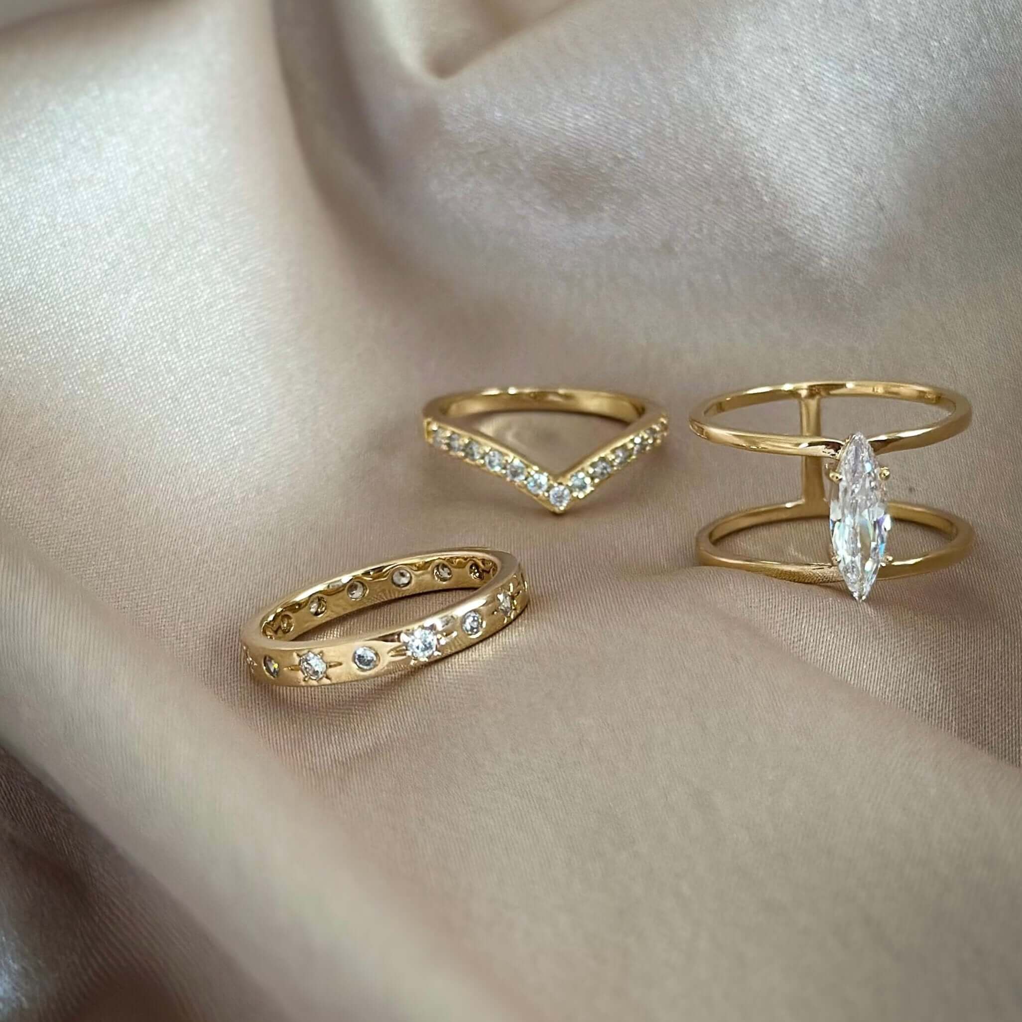 Three Astraea Rings, featuring diamond accents and 18k gold plating, are displayed on beige fabric.