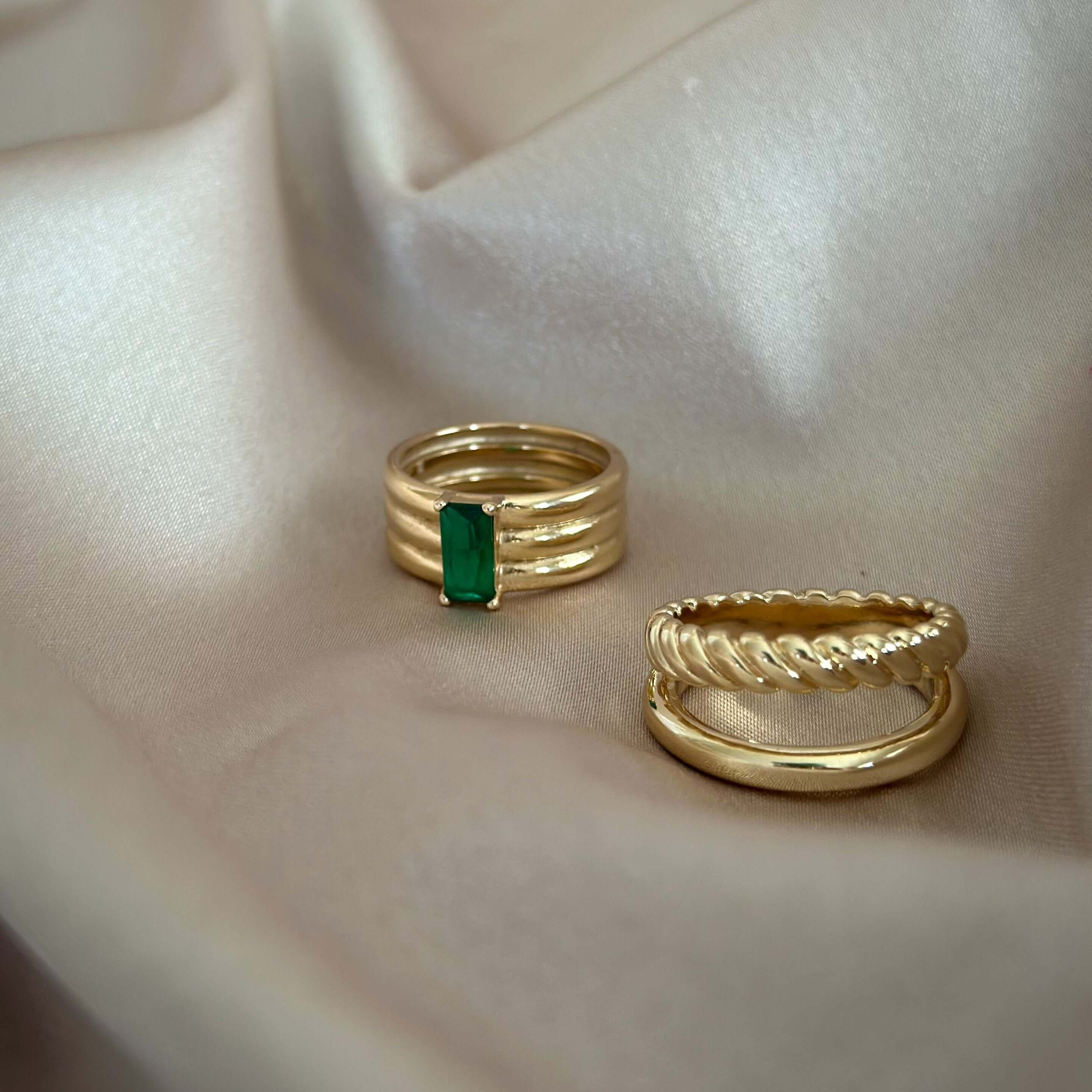 Two gold rings on beige fabric: one ring, the Envy Emerald Ring, features a rectangular green gemstone exuding Emerald Elegance, and the other has a twisted design.