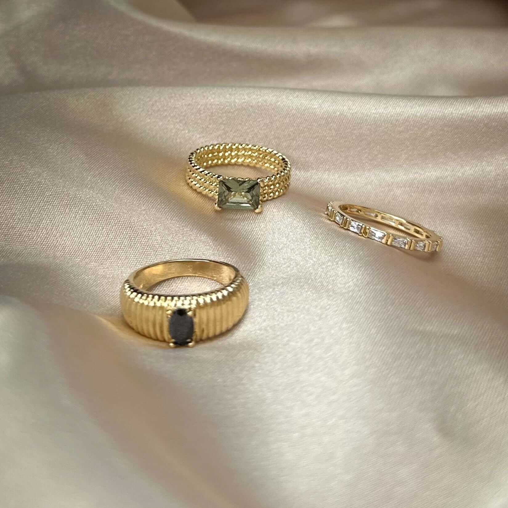 Three Baguette Eternity Bands, featuring 18k gold plating, displayed on a silky beige fabric; each ring boasts unique stone embellishments, epitomizing endless elegance.