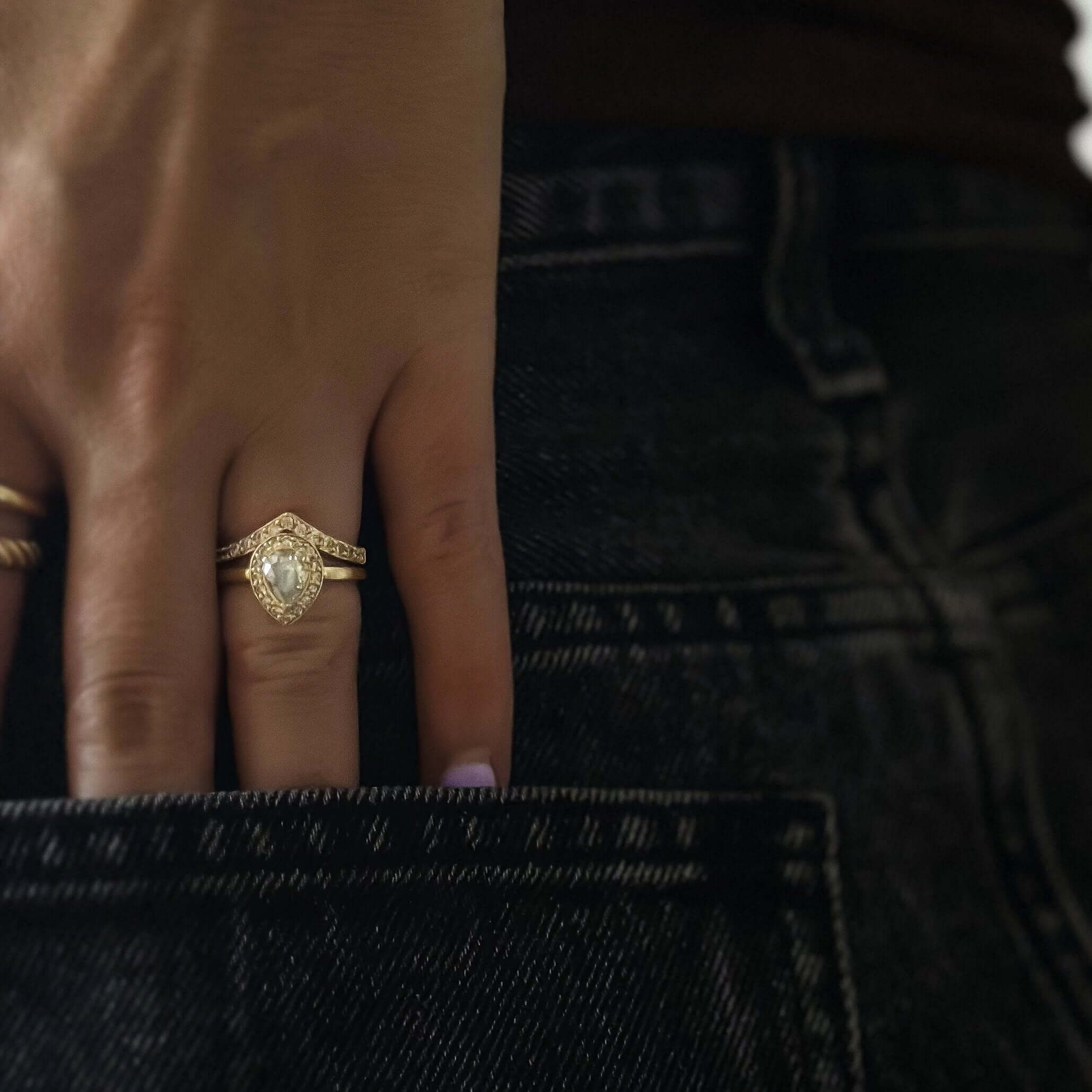 Close-up of a hand with the Pavé Peak Stacking Ring, showcasing its pear-shaped diamond surrounded by pavé diamonds, resting on the back pocket of dark jeans.