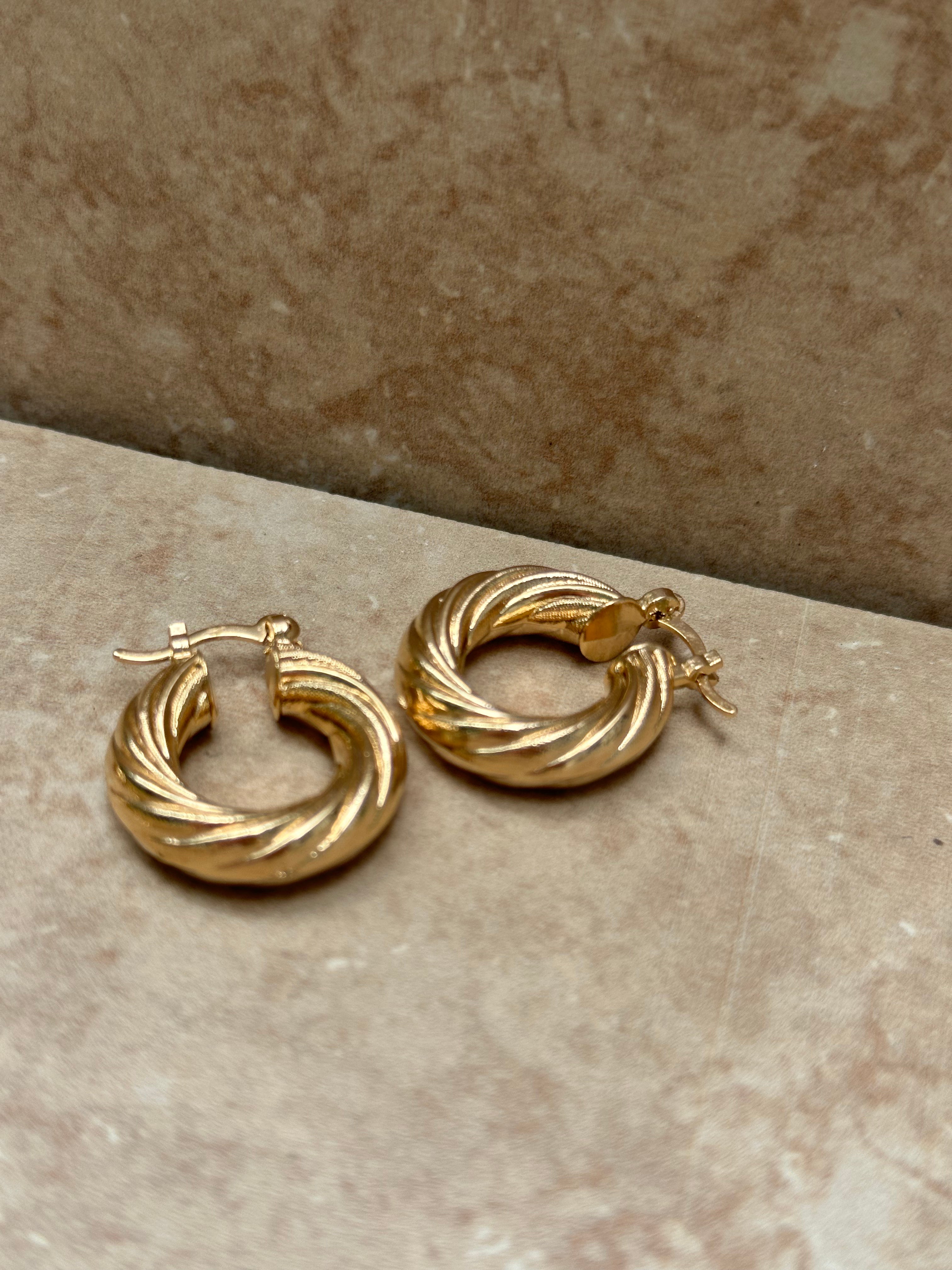 A pair of gold twisted hoop earrings placed on a beige marble surface.