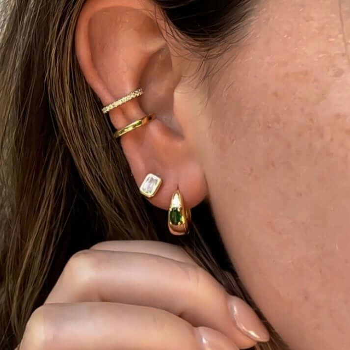 Close-up of a person's ear adorned with multiple gold and diamond earrings, including a hand touching an Envy Emerald Hoop earring. The hoops are complemented by emerald studs, adding a touch of vibrant elegance.