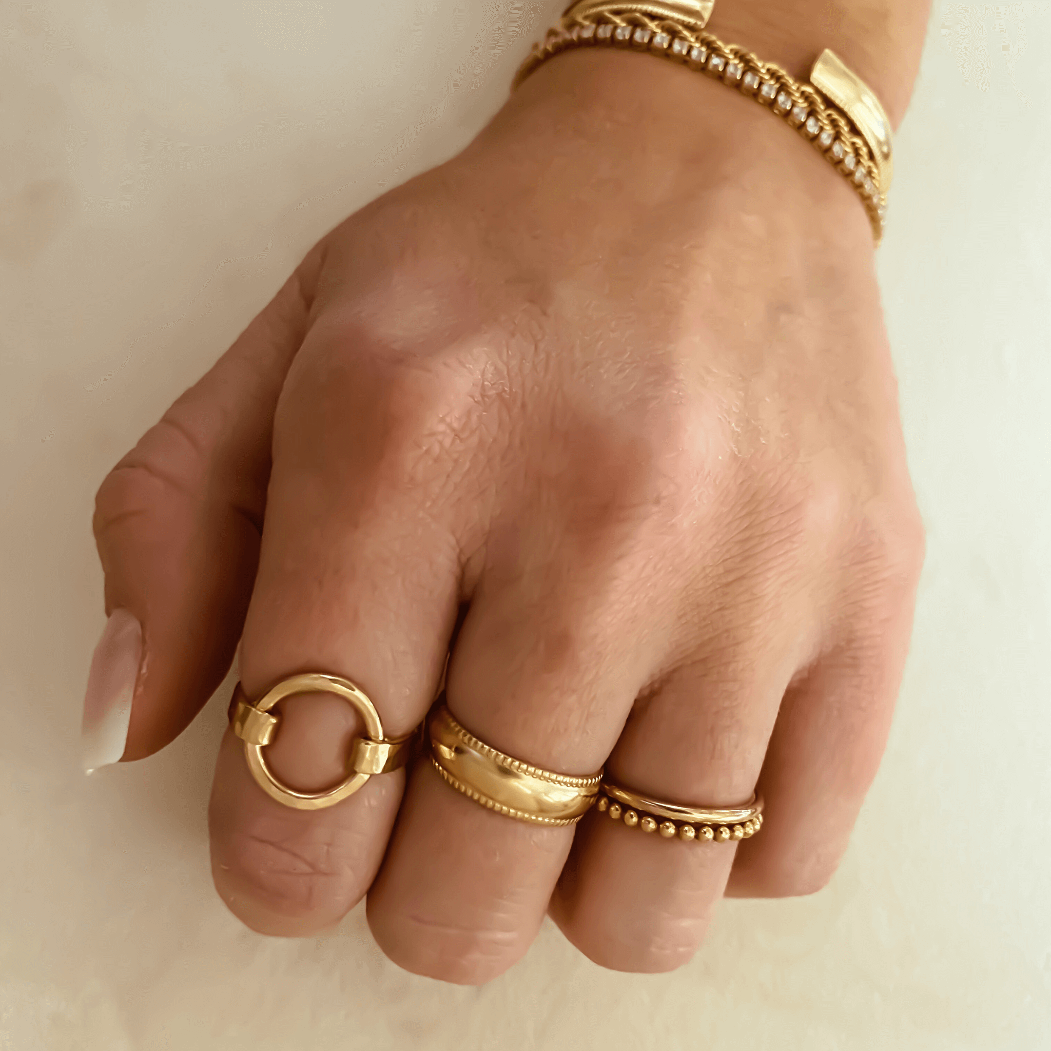 Close-up of a hand adorned with four gold rings, including the elegant Selene Ring in 14k gold fill, and a matching gold bracelet against a light background.