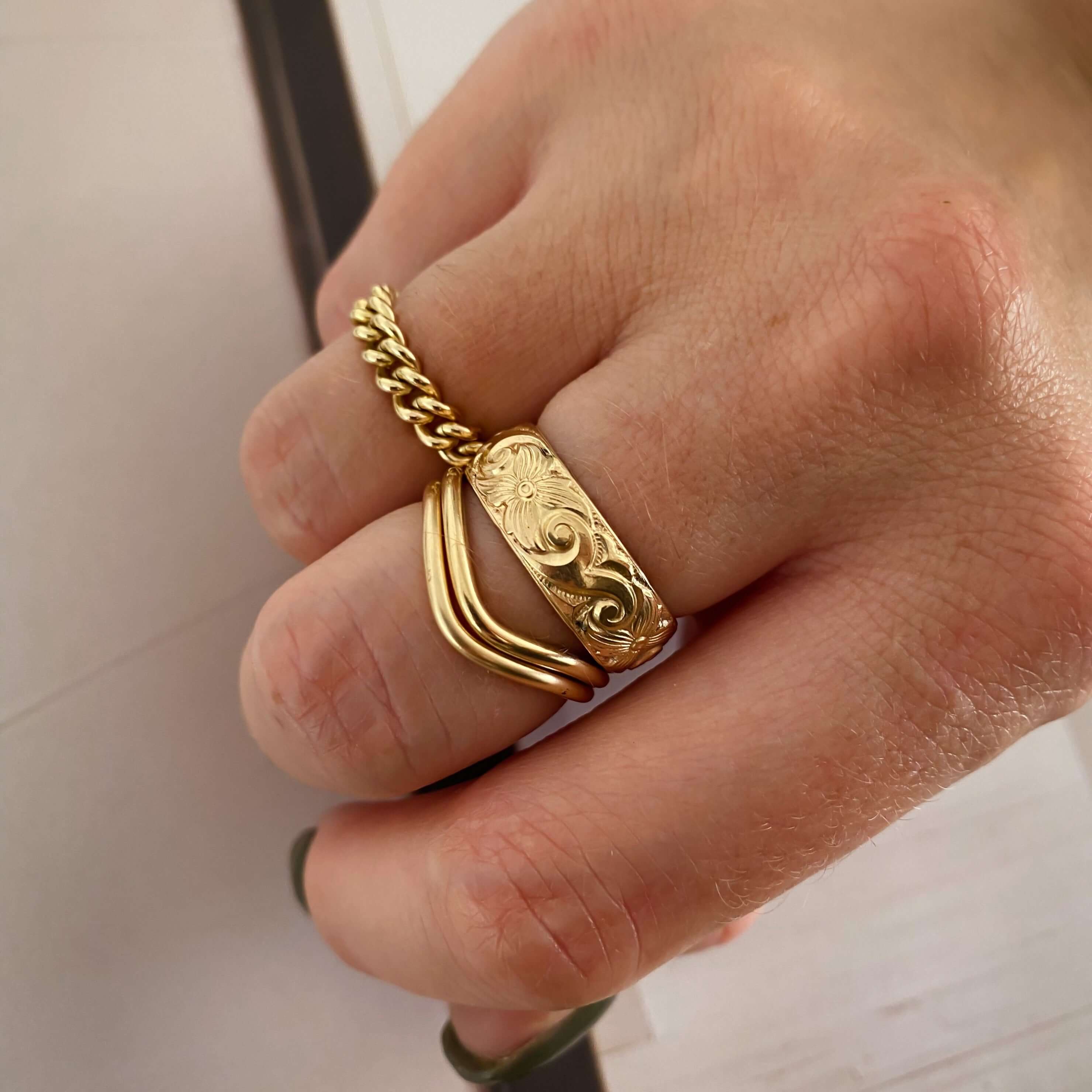 A hand adorned with three unique gold rings: a chain-link ring, a wavy ring, and the stunning Athena Ring featuring an intricate floral design.