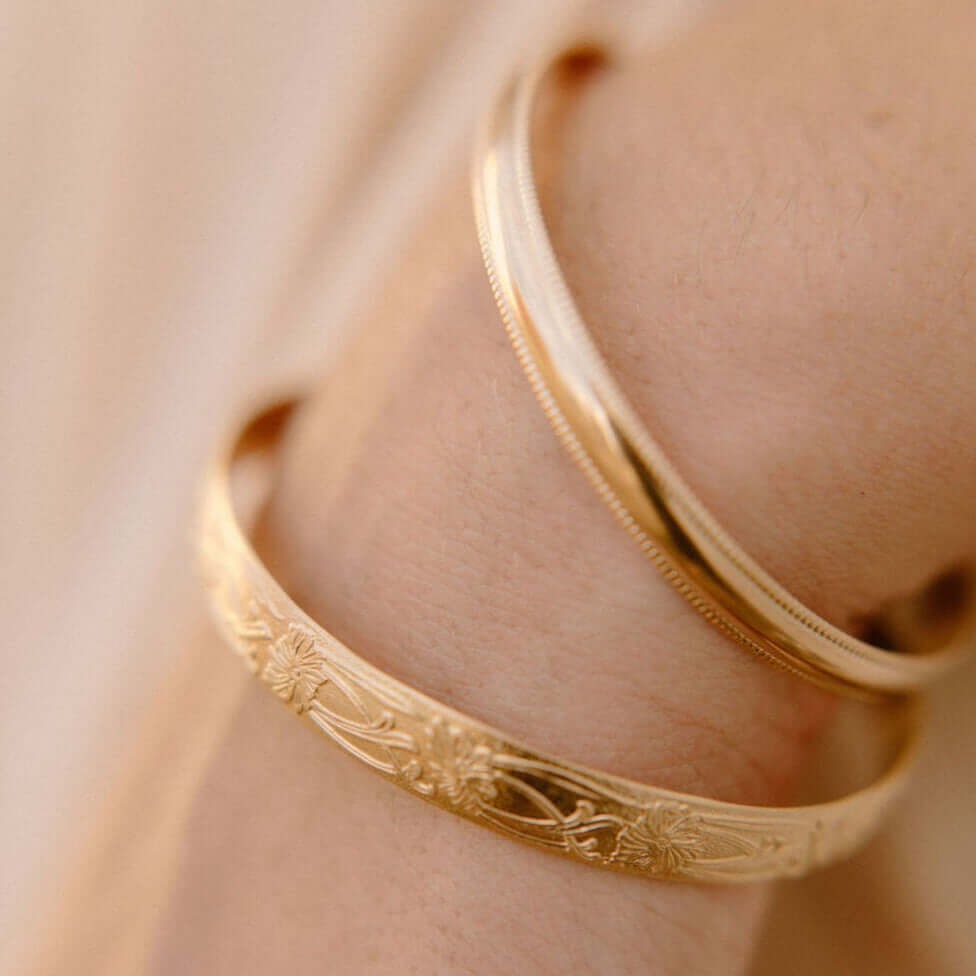 Close-up of two gold bracelets, one smooth and one intricately engraved with floral patterns, worn on a person's wrist.