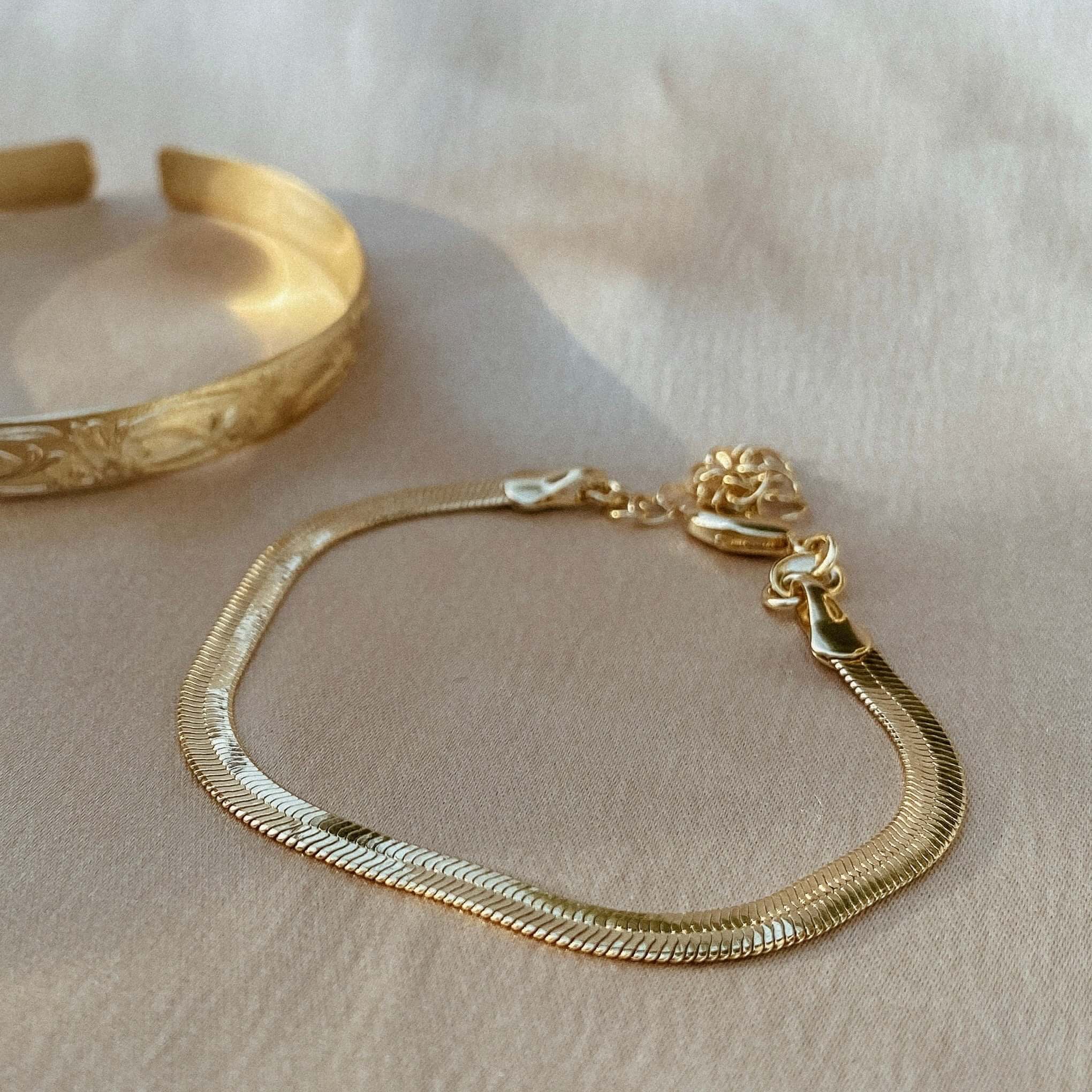 The Bold Herringbone Bracelet, crafted in 18K gold with a lobster clasp, lying on a light surface beside a golden etched bangle.