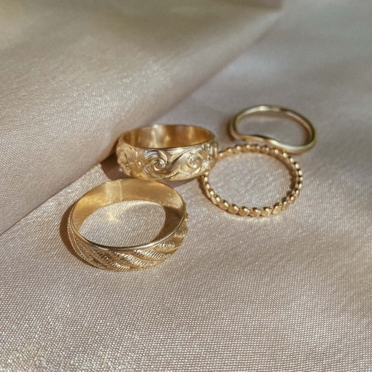 Four gold rings with intricate designs, including the brilliant Athena Ring and a statement band, showcased against a beige fabric backdrop.