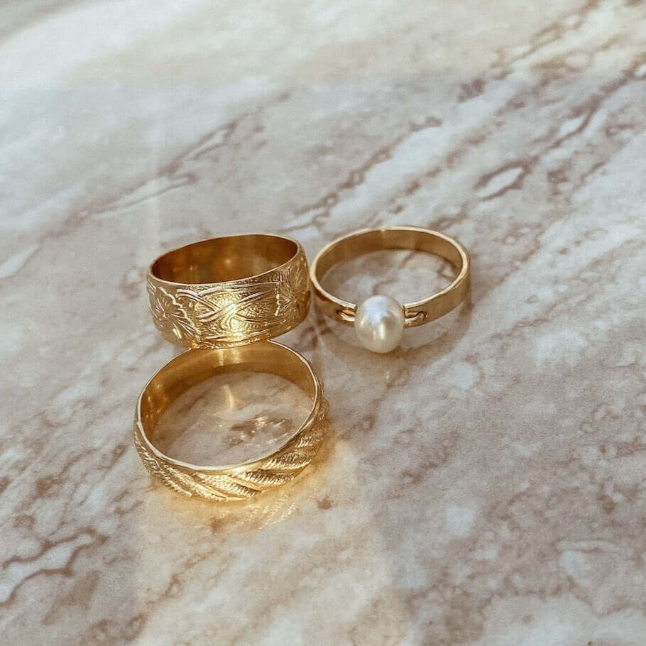 Three gold rings lie on a marble surface, one featuring the Bold Pearl Ring with a stunning freshwater pearl and the others showcasing intricate designs in 14k gold fill. These pieces make for an elegant statement ring collection.