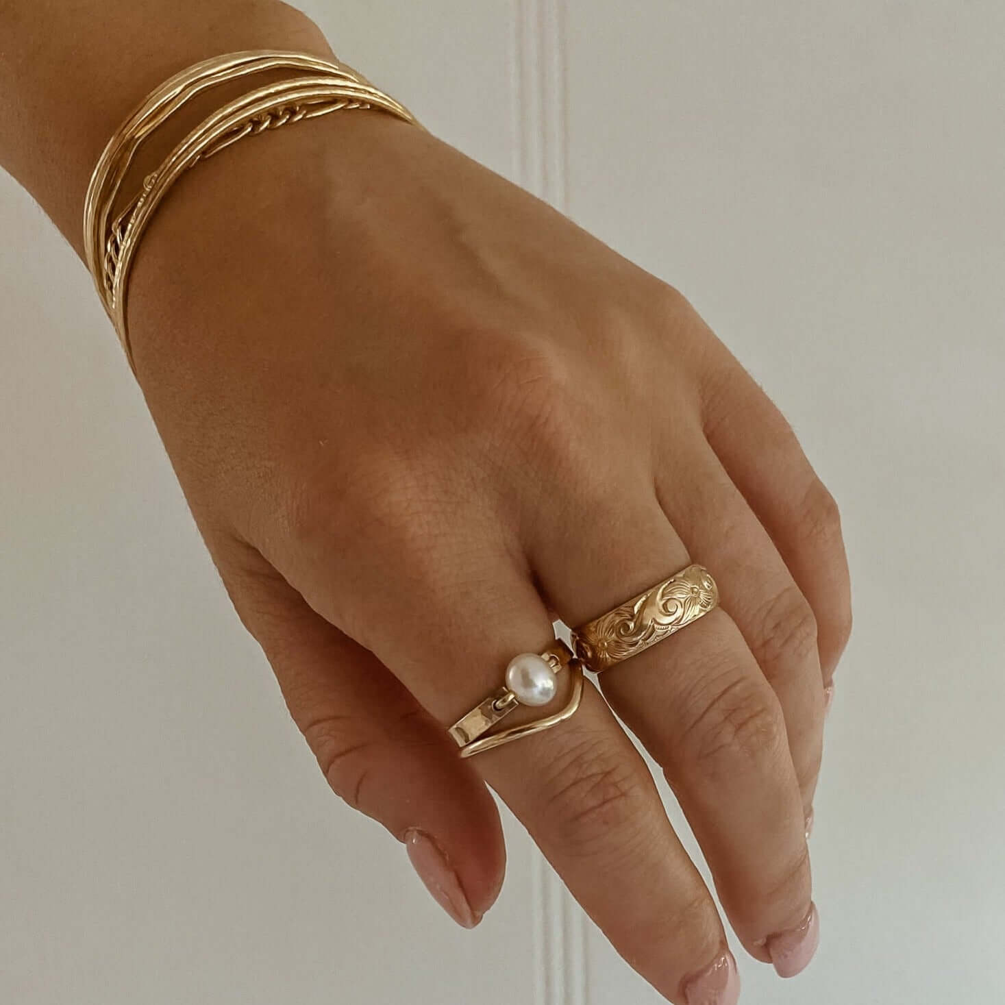 Close-up of a hand adorned with gold rings, including the Athena Ring featuring a pearl, and gold bangles, set against a plain background.