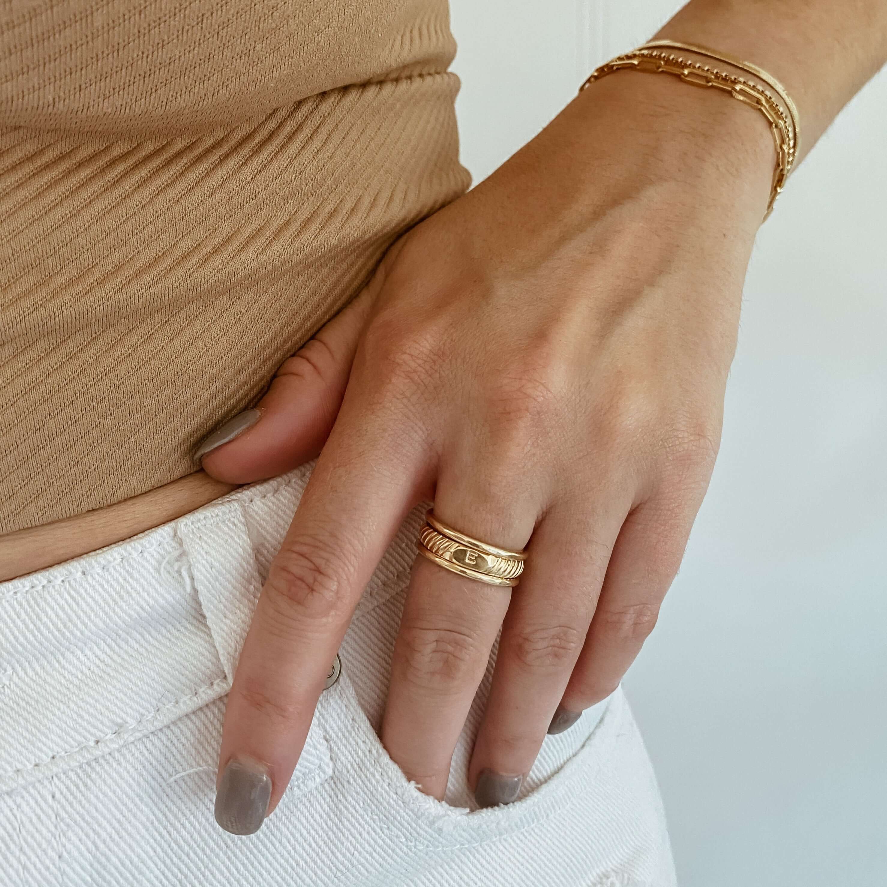 Close-up of a hand touching white jeans, adorned with a personalized Initial Signet Ring in 14k gold fill and a gold bracelet, paired with a beige top.