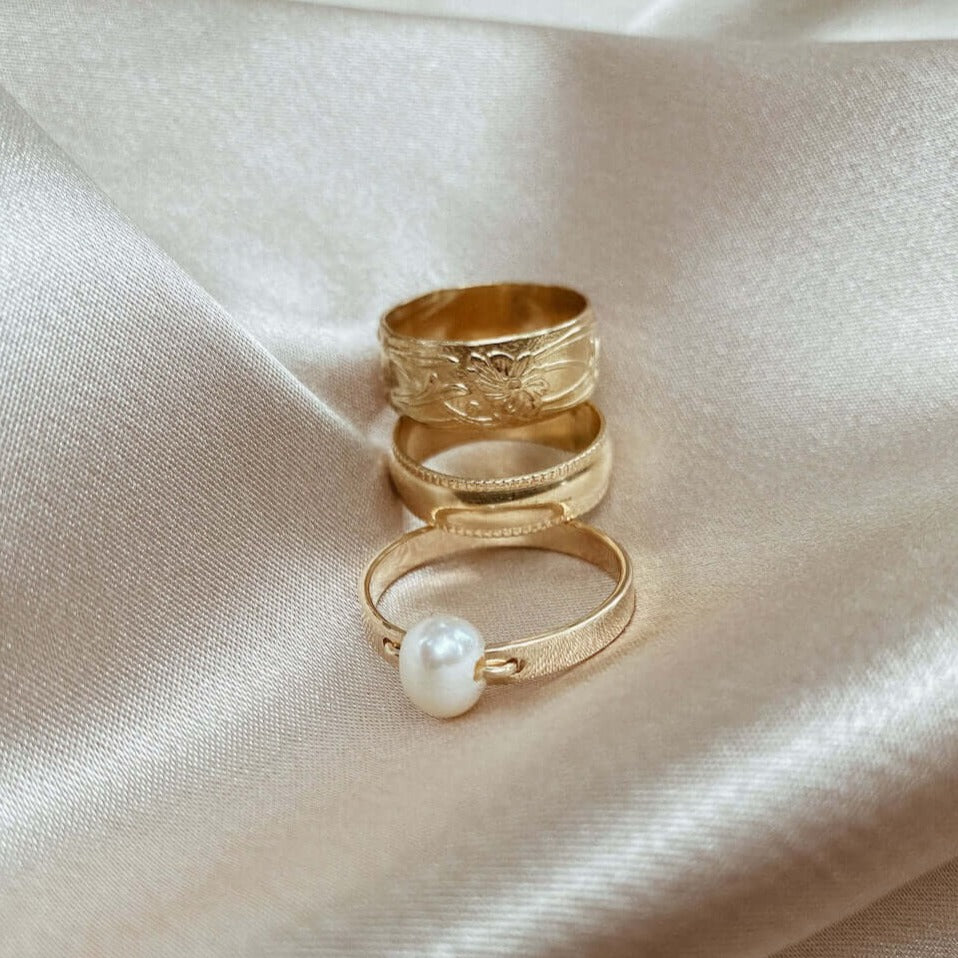 Product Name: Bold Pearl Ring

Sentence: A trio of 14k gold fill rings, featuring one with a textured band, another plain, and the Bold Pearl Ring adorned with a single freshwater pearl, rest elegantly on cream satin fabric.