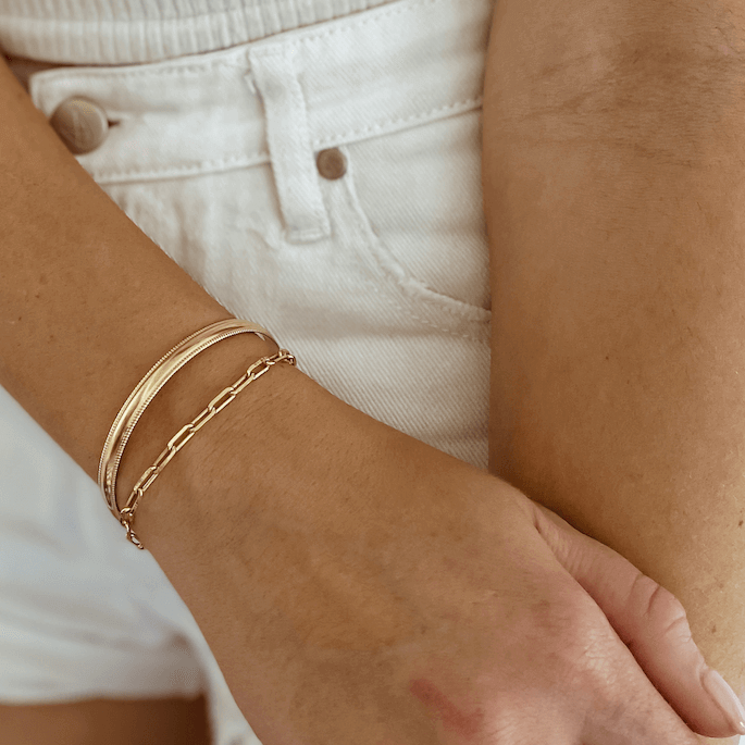 Close-up of a person's arm adorned with an adjustable gold bracelet and the Bold Cuff Bracelet, paired with white high-waisted pants featuring a button design.