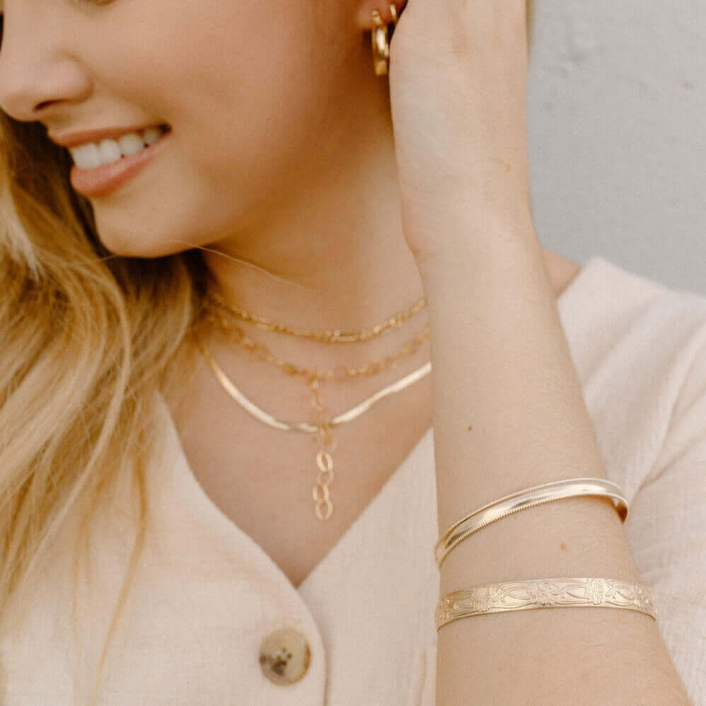 A woman is seen smiling in a beige top, adorned with layered gold necklaces, earrings, and the Bold Cuff Bracelet.