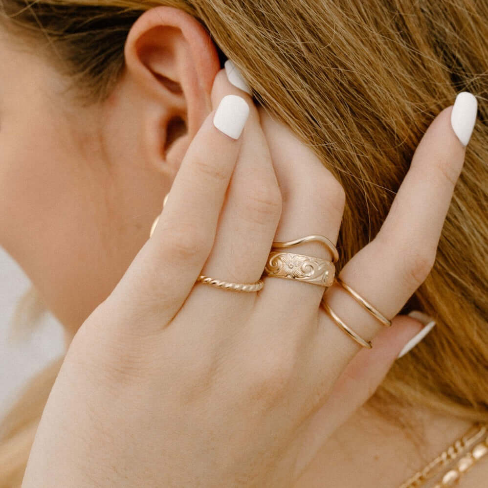 Close-up of a person wearing multiple gold rings, including the striking Athena Ring with its intricate floral design, their hand touching their ear and showcasing white-painted nails.