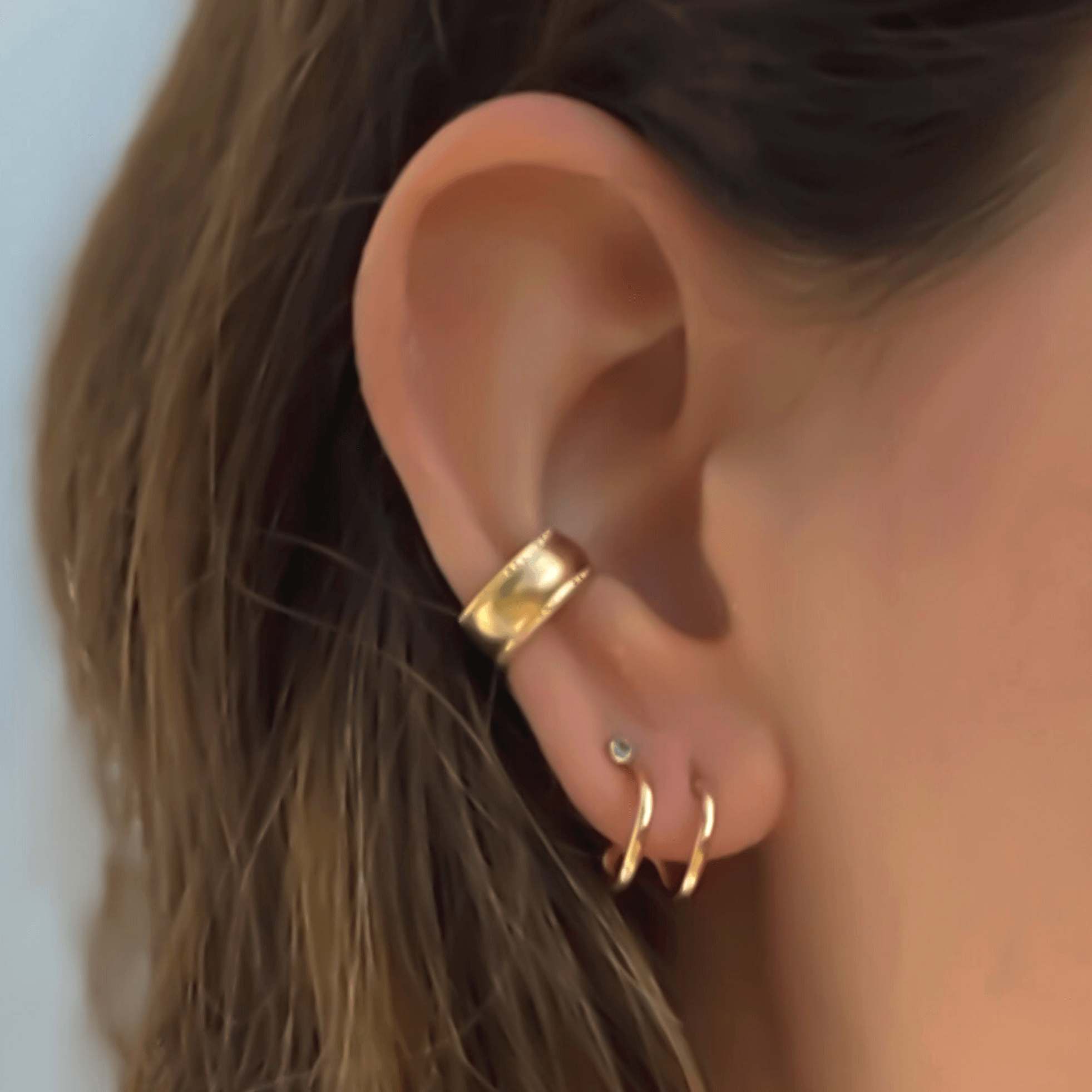 Close-up of a person's ear adorned with three gold earrings: a Bold Ear Cuff at the top, a stud in the middle, and a small hoop at the bottom.